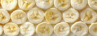 Fresh sliced bananas arranged in rows, showing circular cross-sections with visible seeds, creating a natural yellow pattern on white background.