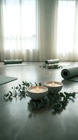 Peaceful yoga studio setup with burning candles, eucalyptus branches and exercise mat on wooden floor near bright windows for meditation and wellness practice.