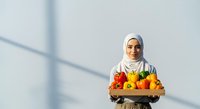 Young woman wearing white hijab holding wooden crate filled with fresh colorful vegetables including bell peppers and tomatoes against white wall with window shadow.