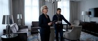 Professional blonde businesswoman and male colleague reviewing documents in modern luxury hotel suite with elegant furnishings and city views.