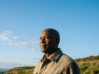 Mature African American man wearing tan jacket standing outdoors with coastal landscape and clear sky behind him, suitable for lifestyle and portrait use.