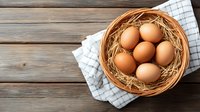 Fresh brown chicken eggs in woven basket with straw nest on rustic wooden table with checkered kitchen towel, top view. Natural organic farm food.