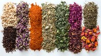 Colorful dried herbs, spices, and tea ingredients arranged in neat rows on white background, including black pepper, rose petals, paprika, herbs, and flower buds for culinary use.