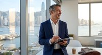 Confident middle-aged Caucasian businessman using tablet in modern office with city skyline view. Professional executive planning strategy during workday.