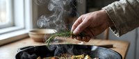 Young adult hand adding fresh rosemary to steaming food in cast iron skillet, cooking preparation with herbs and steam rising, suitable for culinary blogs and food content.