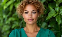 Young mixed race woman with curly blonde hair wearing green medical scrubs smiling confidently against natural green foliage background in outdoor portrait setting.