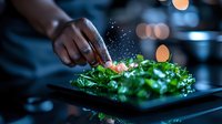 Chef seasoning fresh green salad with salt in modern restaurant kitchen. Water droplets splash dramatically against dark background with bokeh lights.