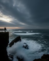 Silhouette of couple standing on dramatic cliff edge watching powerful ocean waves crash against rocky coastline under moody stormy sky at sunset.