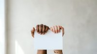 Person with medium brown skin tone holding blank white paper sheet in front of face against neutral wall background for advertising, branding, or promotional messaging purposes.