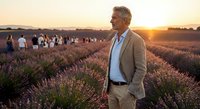 Mature Caucasian businessman in beige suit standing in blooming lavender field at golden hour sunset with tourists in background for relaxation concept.