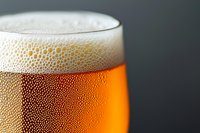 Close-up of cold craft beer glass with condensation droplets and frothy white foam head against dark background, showing amber liquid texture and bubbles.