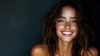 Young mixed race woman with bright white smile and tousled brown hair laughing against dark background, showing natural beauty and joy in lifestyle portrait.