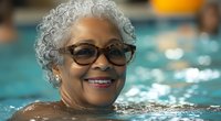 Senior African American woman with silver hair and glasses enjoying water aerobics in swimming pool, displaying genuine happiness and active lifestyle in retirement.