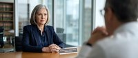 Professional mature businesswoman in navy suit conducting interview or meeting in modern office with city view through windows.