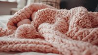 Cozy knitted blanket in trendy coral pink color with braided pattern and pompom detail, soft textured throw creating warm and inviting atmosphere for home decor.