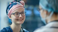 Young female medical professional in glasses and pink surgical cap smiles warmly while conversing with colleague in hospital setting, representing healthcare teamwork.