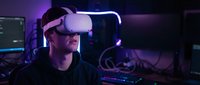 Young Caucasian male gamer wearing VR headset in neon-lit gaming setup with purple and blue ambient lighting for immersive virtual reality experience.