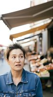 Middle-aged woman of East Asian descent wearing denim jacket standing outdoors near market stall with retractable awnings, expressing surprise or concern. Suitable for lifestyle, retail, and street photography concepts.