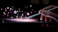 Magical pink glowing particles float above digital tablet screen while finger touches stylus pen in dark environment, creating ethereal technology concept effect.