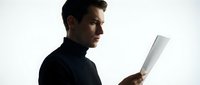 Young man with dark hair and light skin tone, wearing black clothing, holding and examining a tablet device against white background.