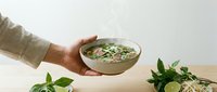 Young adult male hand holding bowl of creamy soup with fresh herbs and vegetables, ideal for food photography, culinary blogs, restaurant menus, and wellness content.