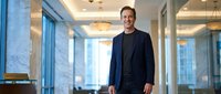 Confident middle-aged Caucasian businessman in dark suit standing in modern corporate office lobby with marble columns and large windows.