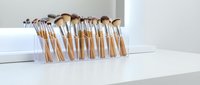 Professional makeup brush set with rose gold handles arranged in clear acrylic organizer on white vanity table for beauty routine and cosmetic application.
