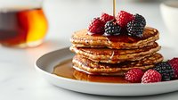 Stack of golden homemade pancakes topped with fresh raspberries, blackberries and flowing maple syrup on white plate against light background. Breakfast setting.