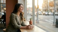 Young professional woman enjoying coffee break at cafe window during golden hour sunset, relaxing after work with warm natural lighting and urban view.