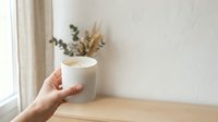 Woman's hand holding white ceramic mug with hot beverage near window, dried flowers in vase on wooden table, morning coffee or tea break at home.