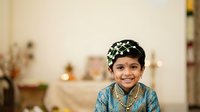 Young South Asian boy wearing traditional blue kurta with embroidered details and decorative headwear, smiling at camera in warmly lit home interior setting.