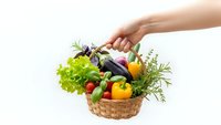Hand holding wicker basket filled with fresh organic vegetables including eggplant, peppers, lettuce and herbs on white background for healthy cooking.