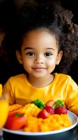 Adorable African American toddler girl in yellow shirt smiling at camera with colorful healthy meal plate featuring fresh fruits and vegetables.
