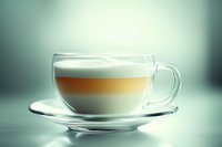 Layered coffee drink with milk foam in transparent glass cup on saucer against blurred turquoise background, showing distinct cream, coffee and milk layers.