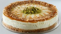 Creamy pistachio cheesecake with caramel drizzle on graham cracker crust, topped with crushed nuts and decorative caramel pattern, served on white plate.