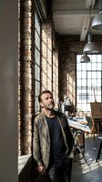 Middle-aged Caucasian businessman in casual blazer standing thoughtfully in industrial loft office with exposed brick walls and large windows.