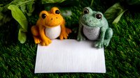 Cute orange and green felt frogs sitting on blank white card against grass background, decorative handmade toys for greeting card or announcement design.