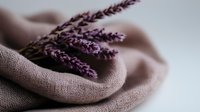 Delicate purple lavender sprigs resting on soft beige linen fabric with gentle folds, creating serene spa and wellness atmosphere. Natural aromatherapy concept.
