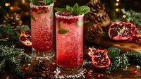 Festive pomegranate cocktails garnished with mint leaves and fresh seeds, served in tall glasses against dark holiday background with pine cones and evergreen decor.