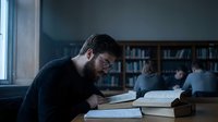 Young bearded man studying intensely with books in university library while students work quietly in background during evening hours.
