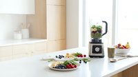 Blender with fresh fruits and greens on a white kitchen counter, surrounded by berries, bananas, and a green smoothie bowl.