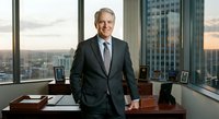 Confident senior Caucasian businessman in dark suit standing in modern corner office with city skyline view at sunset, executive leadership portrait.
