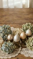 Golden Easter eggs in brass holders with dried hydrangea flowers on vintage lace doily, rustic wooden table background for spring holiday decoration.