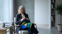 Senior woman with gray hair and light skin tone, wearing dark blazer, sitting indoors holding colorful folded clothes for laundry, domestic chore and household management concept.