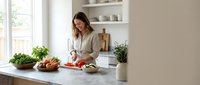 Woman in casual beige shirt preparing fresh vegetables at marble kitchen counter with plants and shelving, ideal for cooking, healthy lifestyle and home interior content.