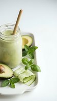 Green smoothie with bamboo straw in mason jar surrounded by fresh avocado, cucumber slices and herbs on white background for healthy lifestyle content.