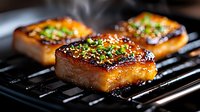 Grilled glazed pork belly slices with caramelized coating and green onion garnish on black grill grates, steam rising in dramatic lighting.
