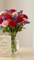 Colorful birthday flower bouquet with red roses, pink carnations and purple statice in glass vase next to greeting card on wooden table for celebration.