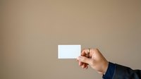 Person with medium skin tone wearing dark blue sleeve holding blank white business card against neutral beige background for corporate branding and professional networking purposes.