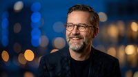 Smiling middle-aged Caucasian businessman with beard and glasses in dark suit jacket against blurred city lights background for corporate headshot.